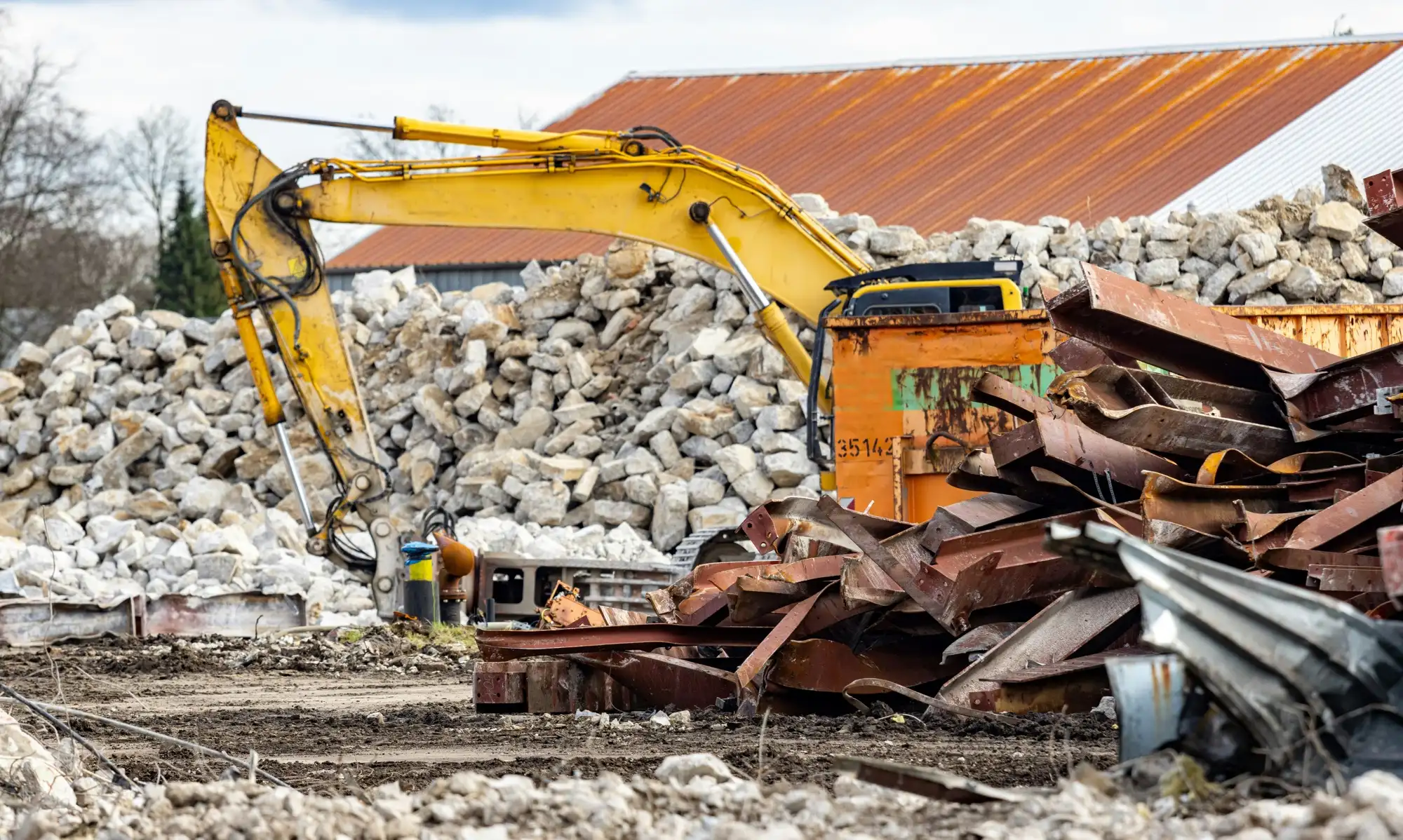 Geltonas ekskavatorius prie išrūšiuotų betono laužo ir metalo krūvų aikštelėje